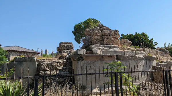 Antik taş kalıntıları, yeşil ağaçlar ve mavi gökyüzü ile çevrili.