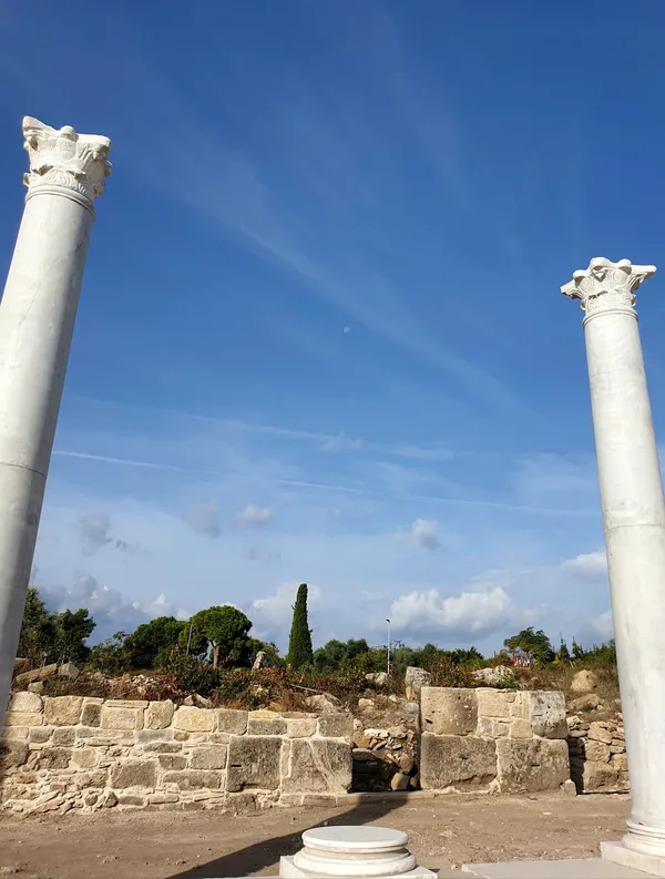 İki mermer sütun, arka planda taş duvar ve yeşil bitkilerle tarihi bir atmosfer sunuyor.