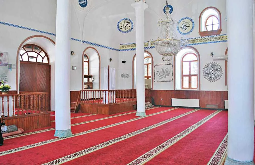 Derin kırmızı halı, beyaz sütunlar ve geleneksel motiflerle süslü cami iç mekanı