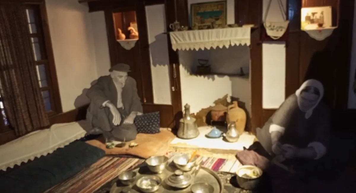 Geleneksel evde yemek hazırlayan kadın ve duvara yaslanmış adam figürü.