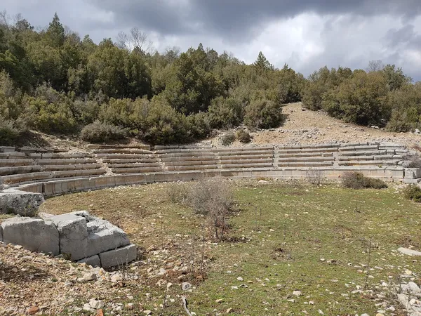 Dağlık alanda yer alan antik tiyatro kalıntısı, taş oturaklar ve yeşil bitki örtüsü ile çevrili.