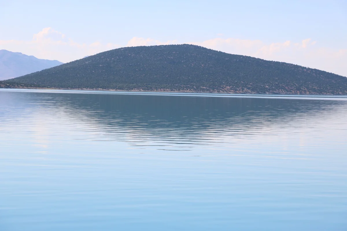 Sakin bir göl manzarası, yansıyan su yüzeyi ve yeşil tepe ile huzur veriyor.