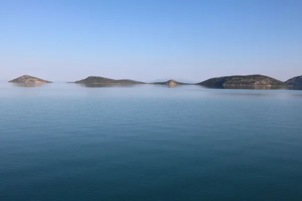 Sakin su üzerinde küçük adalar ve hafif dağlık yapılar, açık mavi gökyüzüyle birleşiyor.