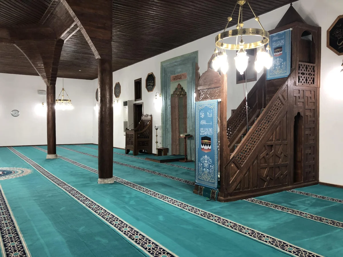 Çeşnigir Paşa Camii iç mekanında yeşil halı, Arapça yazılar ve ahşap minber görünmekte.