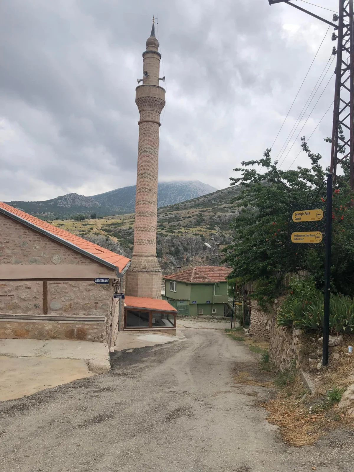 Eski taş yapılar arasında uzun bir minare, yeşil dağlar ve bulutlu bir hava.