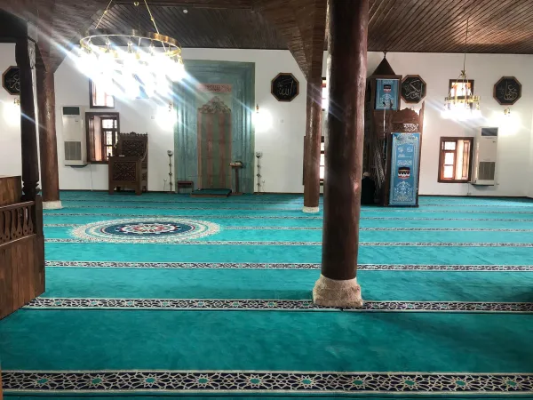 Mavi-yeşil halı, ahşap sütunlar ve klasik İslami motifler ile ferah cami iç mekanı