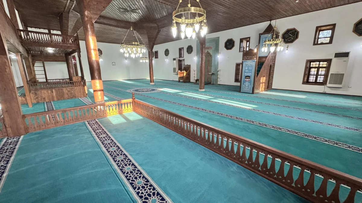 Geniş cami iç mekânı, yeşil zemin, ahşap sütunlar ve doğal ışıkla aydınlatılmış.
