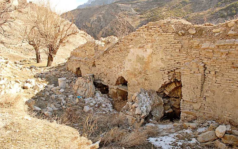 Eskitilmiş yapıda taş ve tuğla duvarlar, yıkılmış kısımlar ve dağlık manzara dikkat çekiyor.