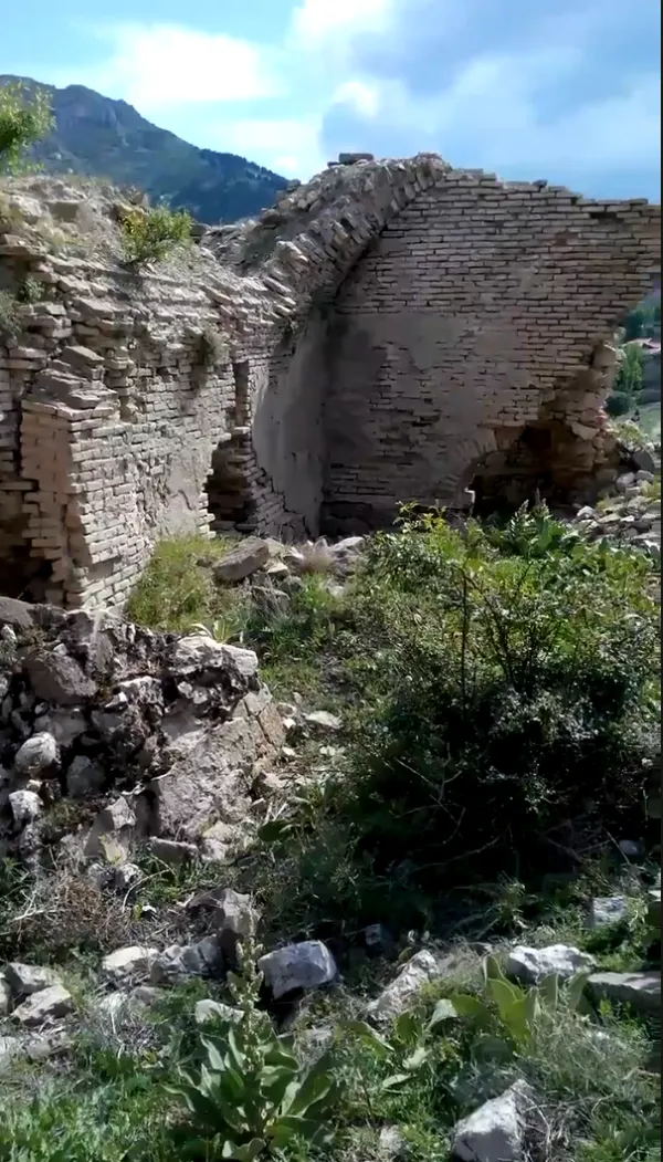 Yıkık bir duvar ve çevresindeki doğal manzara, otlar ve dağlar görünmekte.