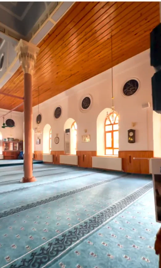 Geniş caminin iç mekanında mavi halı, ahşap tavan ve sarkıt lambalar görülüyor.
