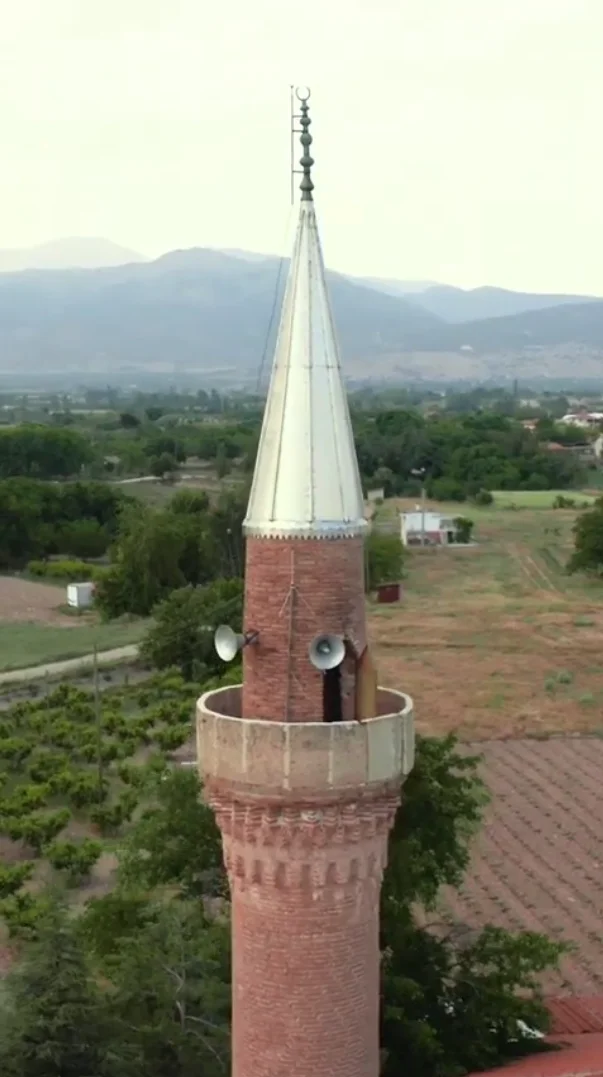 Yeşilliklerle çevrili alanda tuğla minaresi ve metalik şapkasıyla dikkat çeken cami.