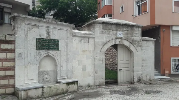 Feyzullah Paşa Camii'nin açık renk taşlarla inşa edilmiş girişi, yuvarlak kemerli ahşap kapı ve sol tarafındaki çeşme.