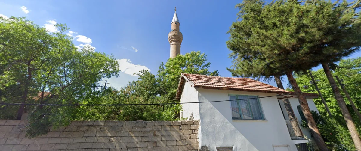 Mavi gökyüzü altında yüksek bir minare ve çevresinde yeşil ağaçlar ile beyaz bir ev