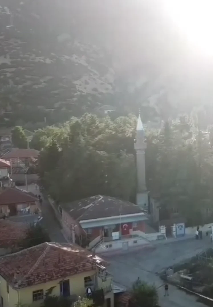 Küçük bir köyde cami minaresi, Türk bayrağı ve ağaçlarla çevrili dağlar.