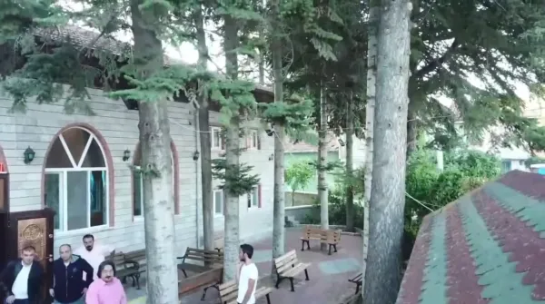 Geniş bir alanda ahşap cami, çam ağaçları ve oturan insanlar.