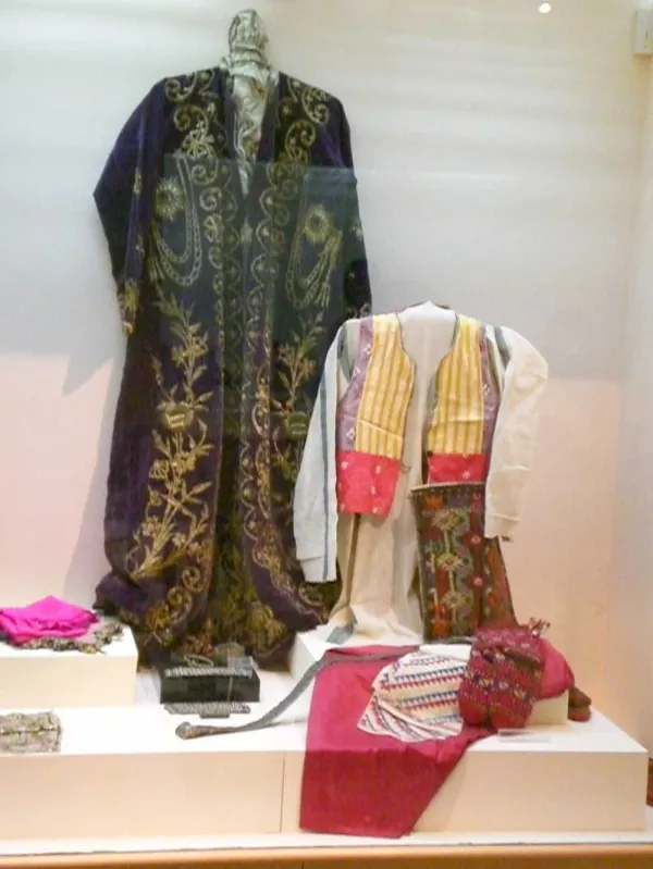 Geleneksel kıyafetlerin sergilendiği vitrin, mor elbise, beyaz gömlek ve renkli desenli aksesuarlar.