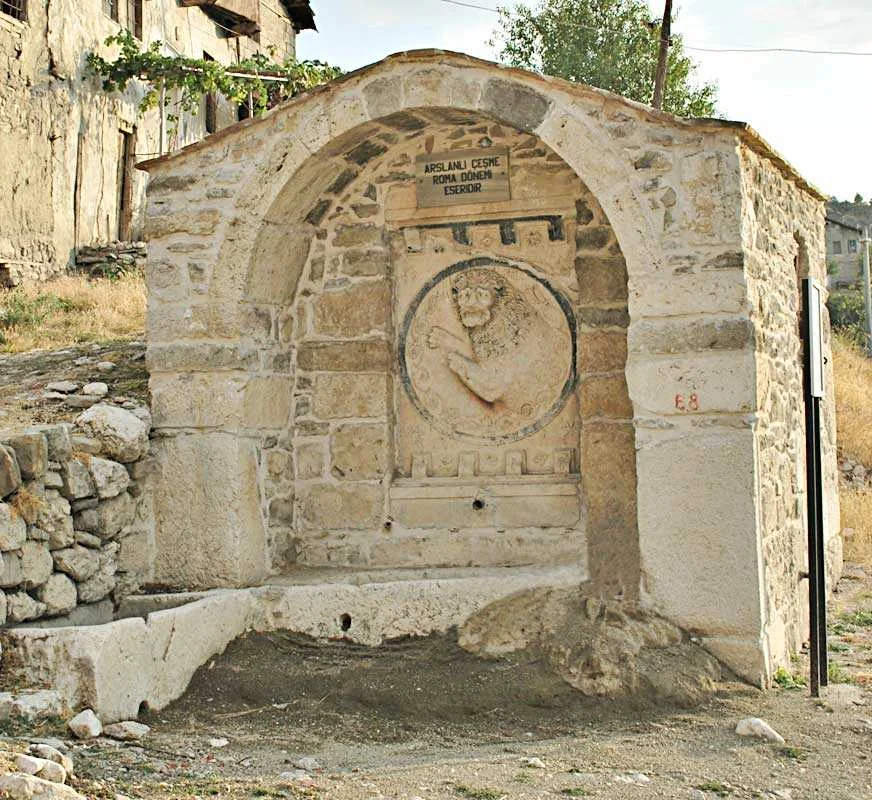 Taşlardan yapılmış tarihi çeşme, üstünde aslan kabartması ve yeşil alanlar ile çevrili.