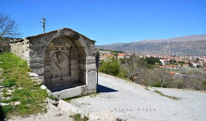Taş aslanlı çeşme, yeşil bitkiler ve dağlarla çevrili, mavi gökyüzü altında.