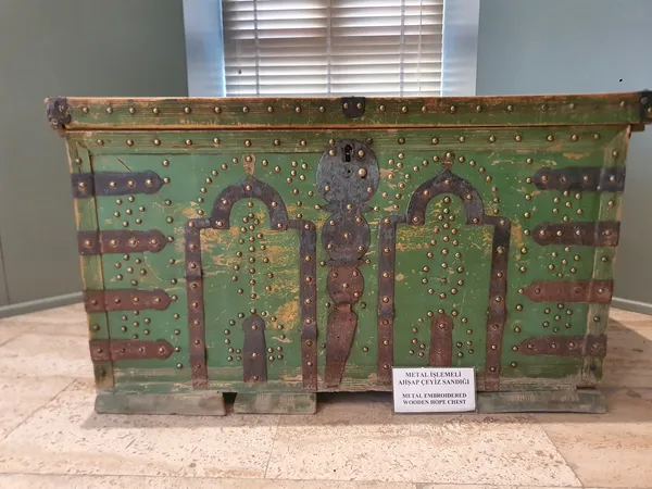 Yeşil metal işlemeli ahşap çeyiz sandığı, önünde bilgi kartıyla sergileniyor.