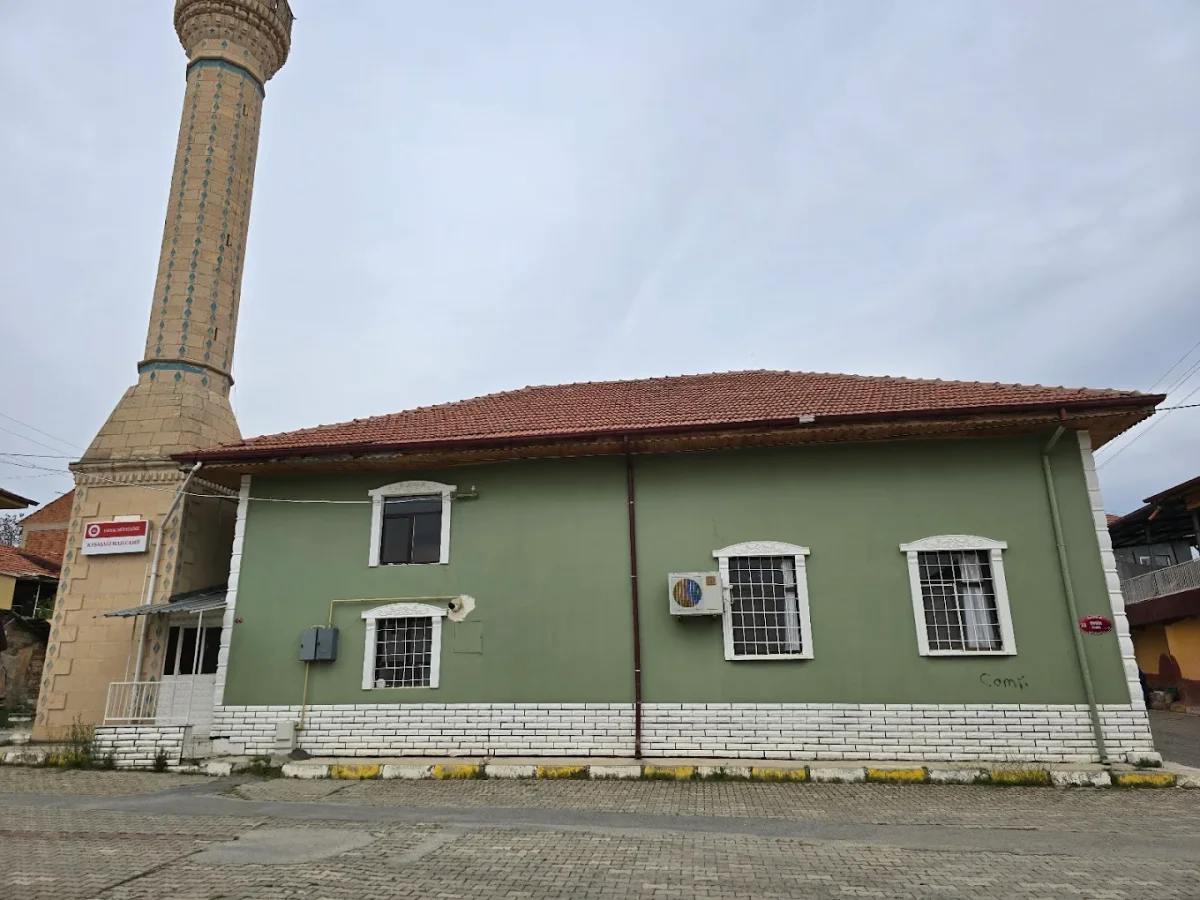 Yeşil duvarlı tek katlı cami ve yüksek taş minaresi, bulutlu gökyüzü arka planda.