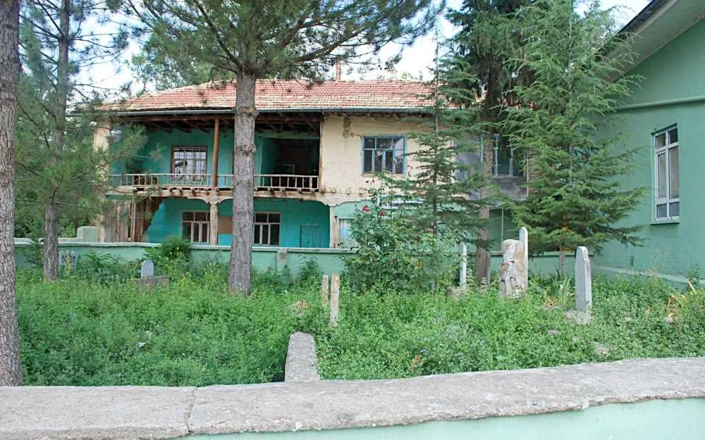 İki katlı eski bina, yeşil-mavi tonlarda, mezar taşları ve yoğun ağaçlarla çevrili bahçede.