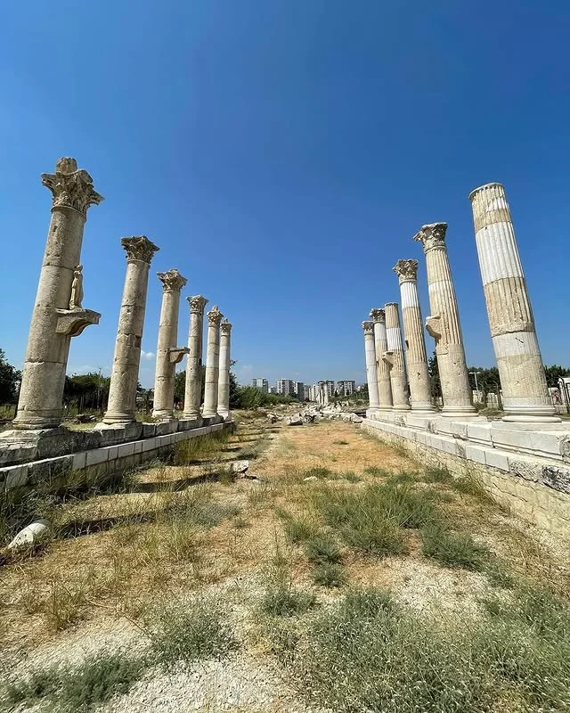 Büyüleyici Sütunlar - Antik ve Modernin Dansı! Antik kalıntılara ait sütunlar, bazıları ayakta bazıları hasar görmüş, arka planda modern binalar var.