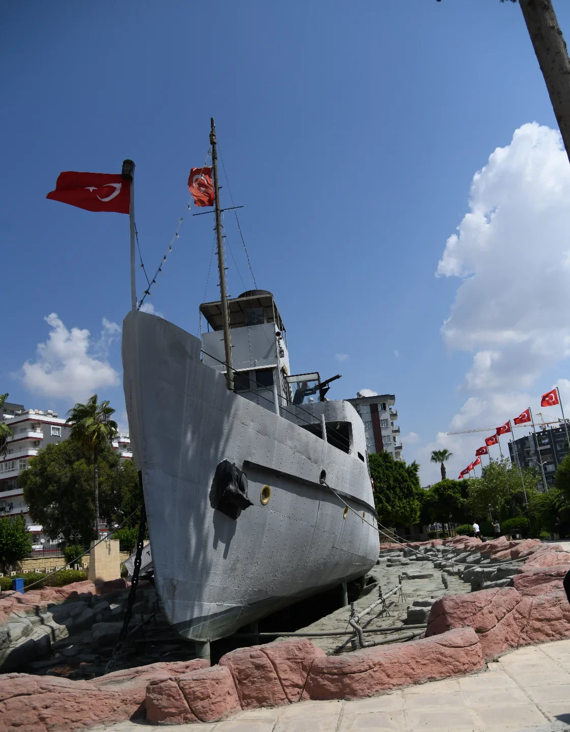 Nusrat Mayın Gemisi - Tarihin Derinliklerine Yolculuk Büyük bir gemi, Türk bayrakları ile karaya yerleştirilmiş, çevresinde parke taşları döşenmiş yol var.
