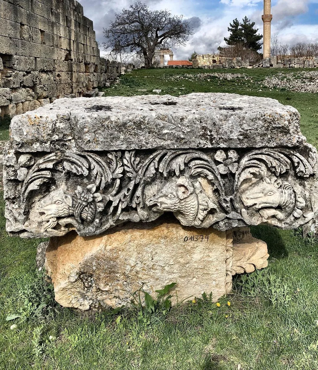 Uzuncaburç'taki Antik Sütun Başlığı İşlemeli sütun başlığı üzerinde aslan ve keçi figürleri, yeşil alanda taş yapılar ve ağaçlar ile dramatik gök