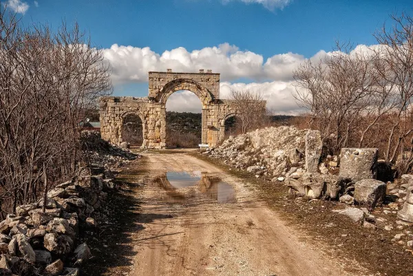 Taşlardan yapılmış kemerli kapı kalıntısı, mavi gökyüzü ve sulama yapan su birikintisi ile çevrili.