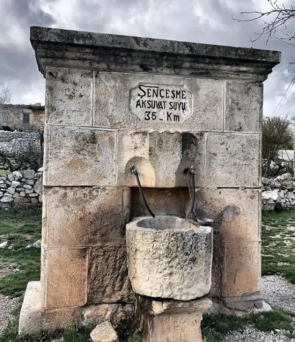 Taş yapıdan bir su kaynağı, önünde musluklar ve doğal peyzaj.
