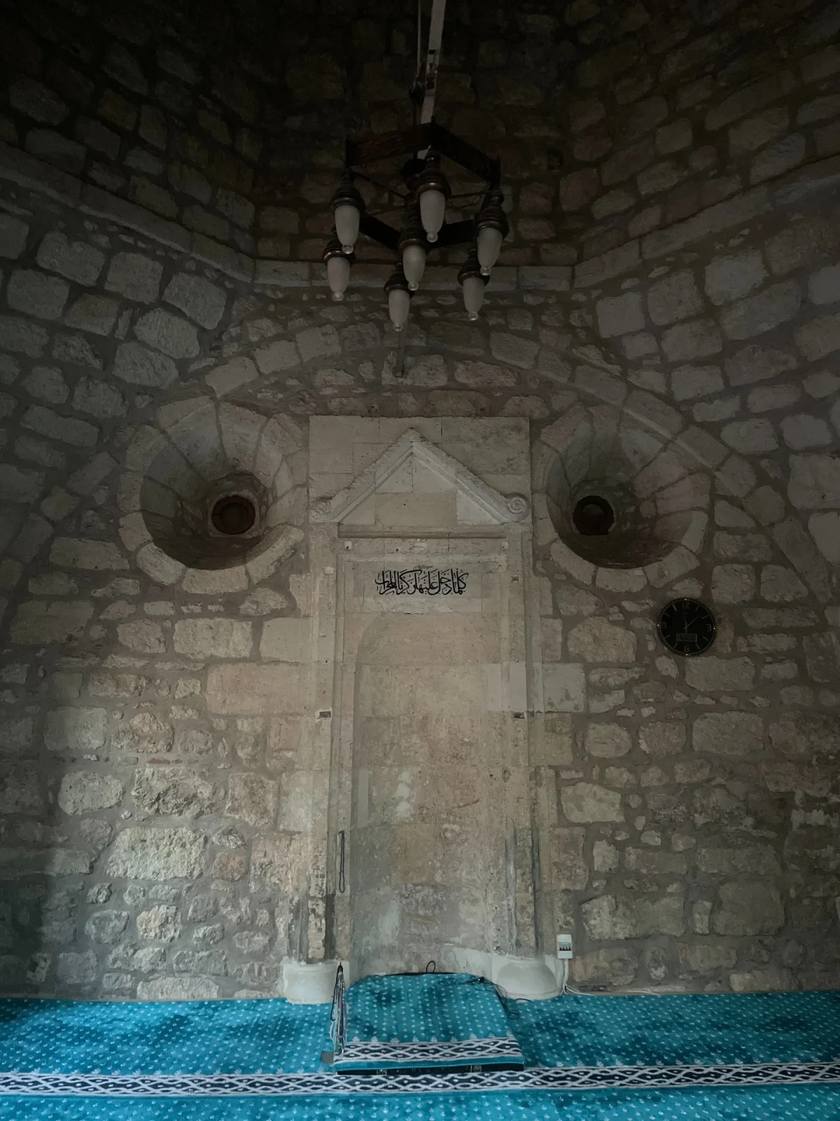Tarihi Atmosferin Mistik Havası - Arapça Yazılı Kapı ve Mavi Halı! Taş duvarlarla çevrili, loş ışıklı tarihi mekan; Arapça yazılı kapı ve mavi halı ile mistik bir ortam.