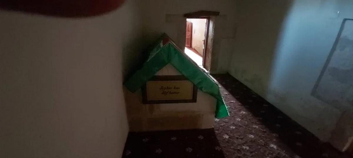Hüzünlü Bir Mekan - Yeşil Örtüyle Kaplı Mezar Yeşil örtüyle kaplı mezar, sade duvarlar ve halılı zeminle iç mekan görünümü.