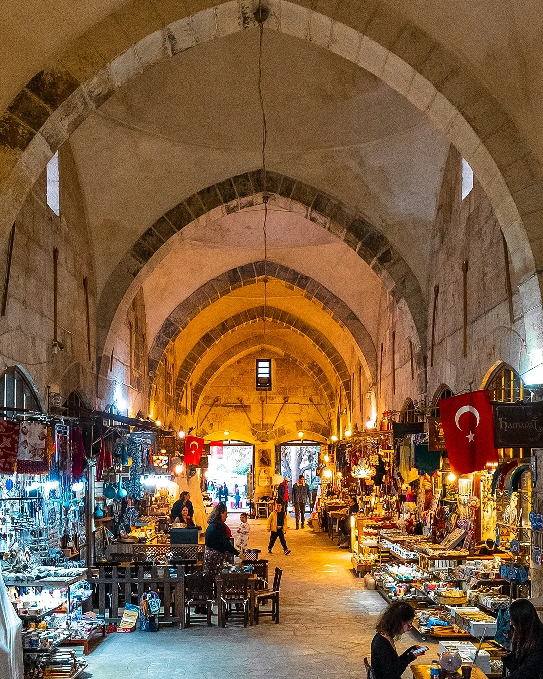 Tarihi Çarşının Büyüsü - Hediyelik eşyalar, alışveriş yapanlar ve Türkiye bayrağı Tarihi çarşıda hediyelik eşyalarla dolu tezgahlar, alışveriş yapan insanlar ve Türkiye bayrağı görünmektedir.