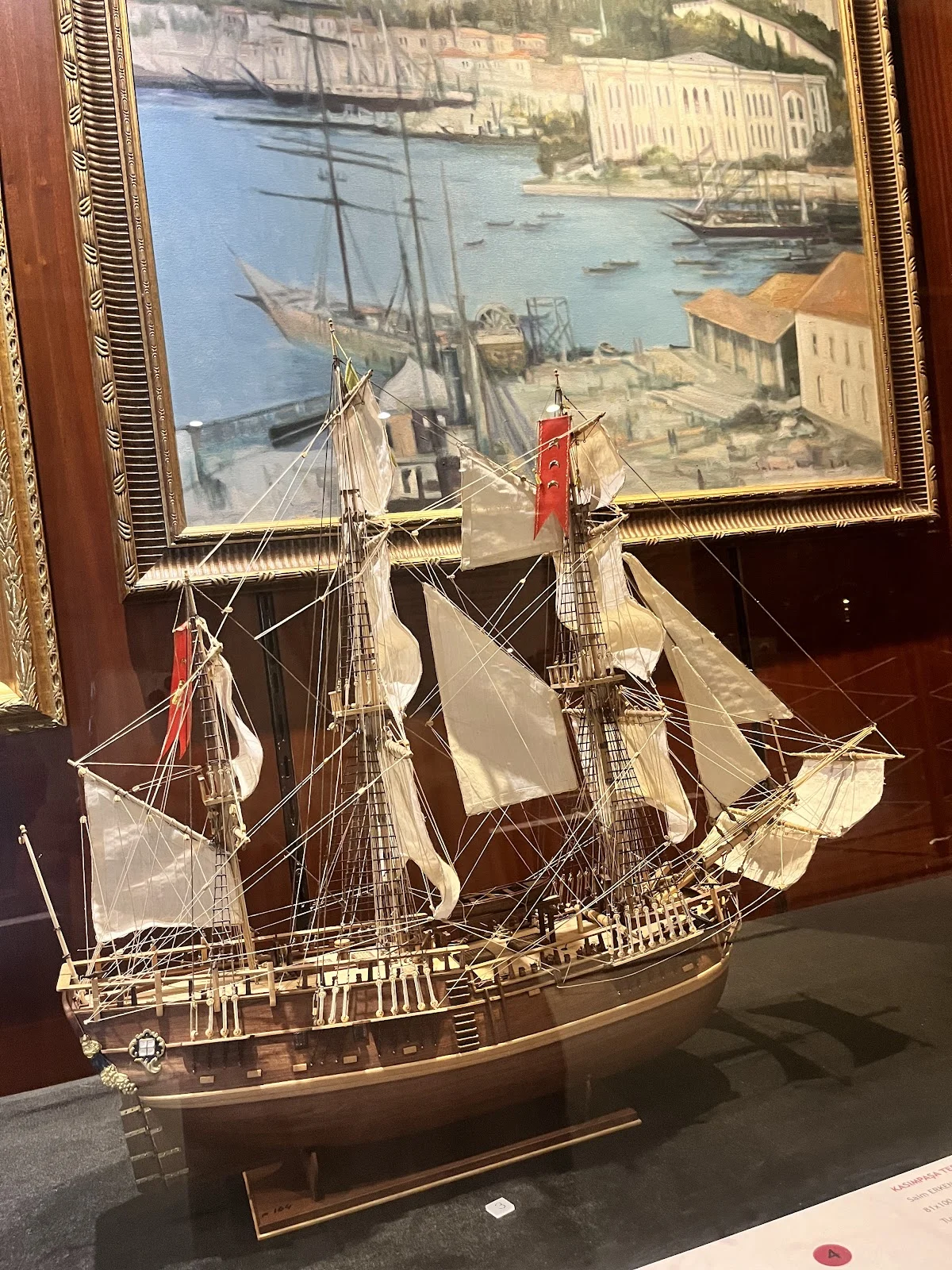 Ahşap Gemi Maketi ve Deniz Manzarası - Tarihi Yolculuk! Ahşap gemi maketi, açılmış beyaz yelkenlerle vitrine yerleştirilmiş, arka planda deniz manzaralı tablo