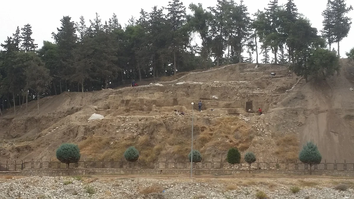Yumuktepe: Tarih ve Doğa - Arkeolojik Kazıların Yeşille Buluşması! Arkeolojik kazı alanında işçiler, taş duvar kalıntıları ve yeşil ağaçlar.