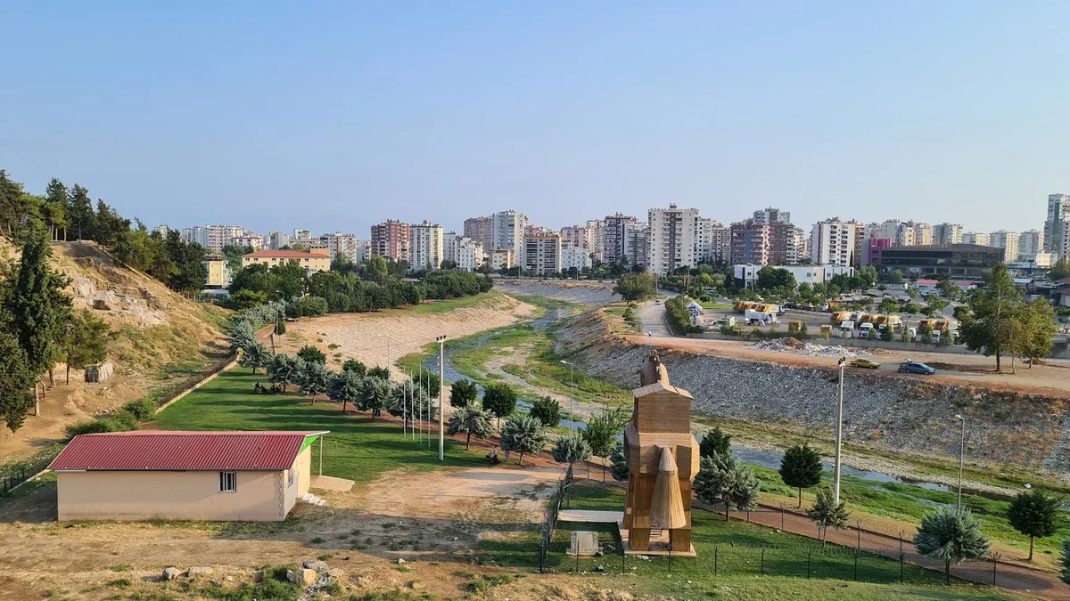 Yumuktepe: Doğa ve Şehir - Yeşil Alanlar ve Heykelin Şehirle Buluşması! Yeşil alan, ağaçlar ve ilginç heykel ile şehir silueti.