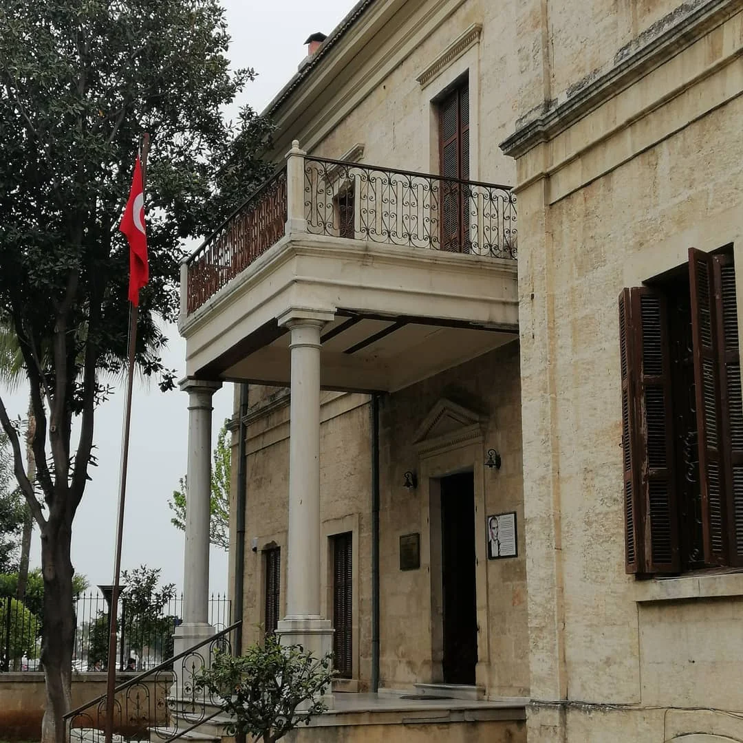Mersin Atatürk Evi'nin Tarihi Mimari Güzelliği: Sanatın ve Mimarinin Buluştuğu Nokta! Mersin Atatürk Evi'nin cephe görünümü, balkon ve Türk bayrağı ile çevre ağaçlar