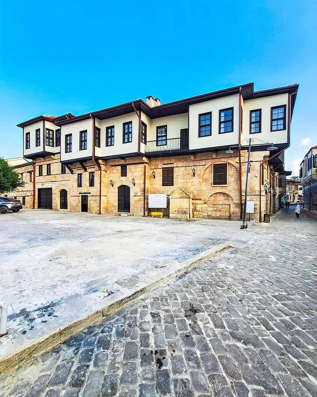 Tarihi Tarsus Evleri - Geçmişin İzleriyle Buluşma! Kültürel Mirasın Gizemlerini Keşfetmeye Hazır mısınız? Tarihi iki katlı taş bina, beyaz cephe ve ahşap detaylarla çevrili açık alan