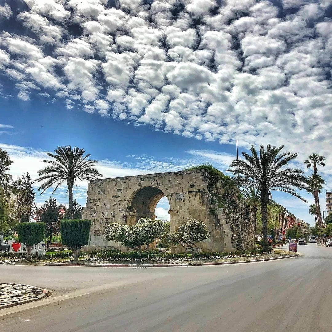 Kleopatra Kapısı: Tarihin Kapısını Aralayın Yeşilliklerle çevrili büyük taş kemer, mavi gökyüzü ve palmiye ağaçları ile turistik bir atmosfer sunuyor.
