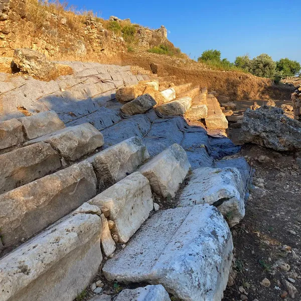 Geniş taş döşemeli alan ve arka planda duvar kalıntılarıyla doğa manzarası