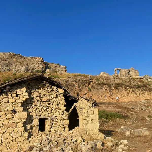 Taş yapılarla inşa edilmiş eski bir ev, arka planda antik kalıntılarla birlikte doğal bir manzara.
