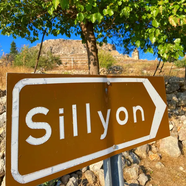 Sillyon yönüne işaret eden kahverengi tabela, arka planda tarihi kalıntılarla birlikte yeşil ağaçlar.