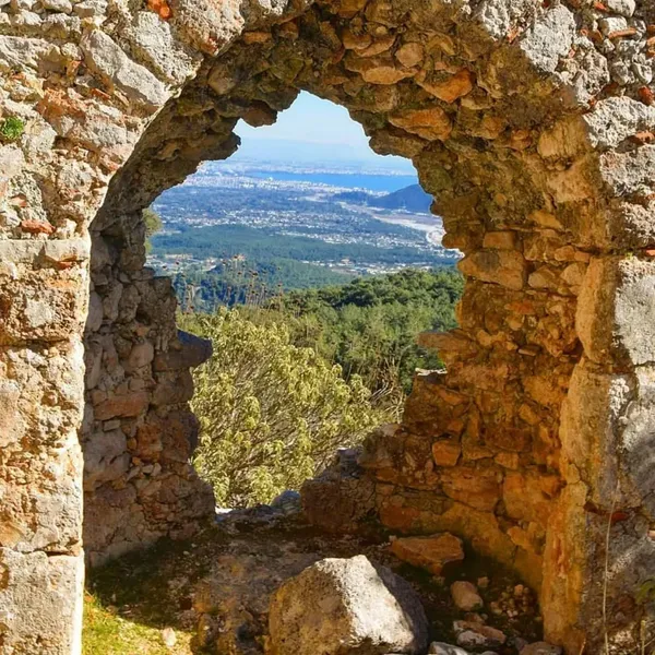 Taş yapıda tarihi pencere, yeşil ormanlar ve kıyı manzarasıyla aydınlatılıyor.