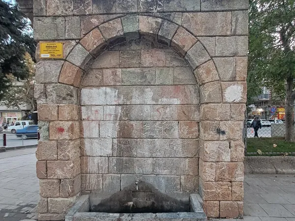 Taşlardan inşa edilmiş bir çeşme, su musluğu ve çevresindeki ağaçlar.