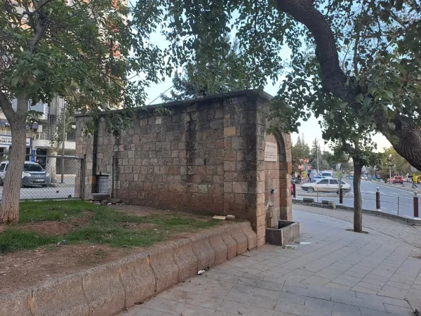 Taş duvarla çevrili alanda yer alan çeşme, etrafındaki ağaçlar ve şehir manzarasıyla dikkat çekiyor.