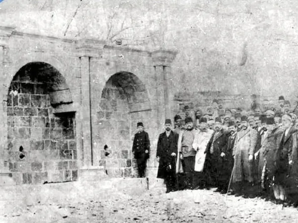 İki taş kemerin önünde resmi giyimli bir grup insan, arka planda yapısal kalıntılar görünüyor.