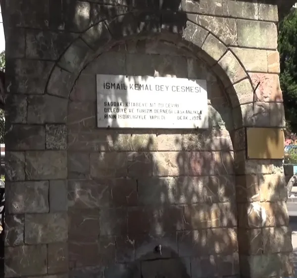 Tarihi taş çeşme, üstünde plaka ve yeşil bitki örtüsüyle çevrili.