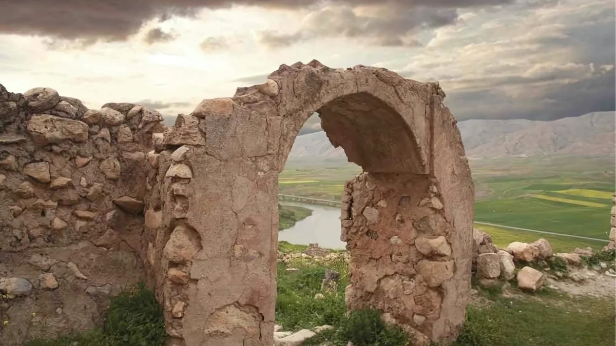 Yıkık kemerin arkasında yeşil alan ve nehir manzarası, gri bulutlu gökyüzüyle doğal bir atmosfer sunuyor.
